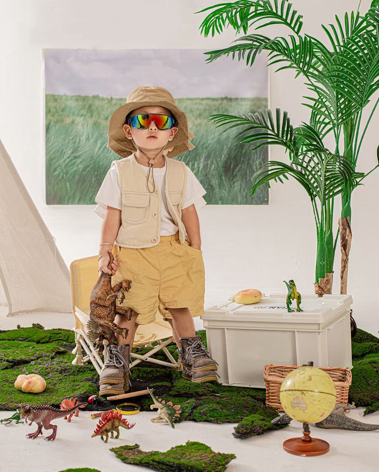 A child in an explorer outfit with colorful sunglasses, hat, and vest, holding a toy dinosaur. The scene includes a mossy ground, toy dinosaurs, a globe, and a palm plant, with a nature painting in the background.